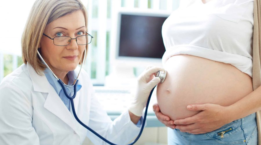 Mature gynecologist looking at camera while examining the belly of a pregnant young woman