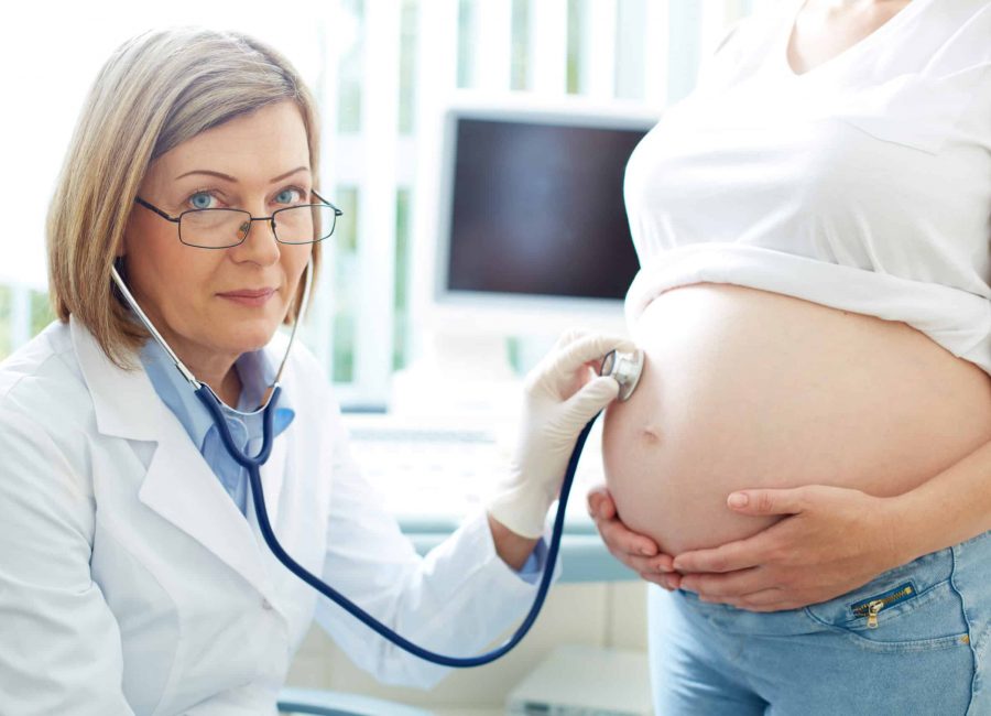 Mature gynecologist looking at camera while examining the belly of a pregnant young woman